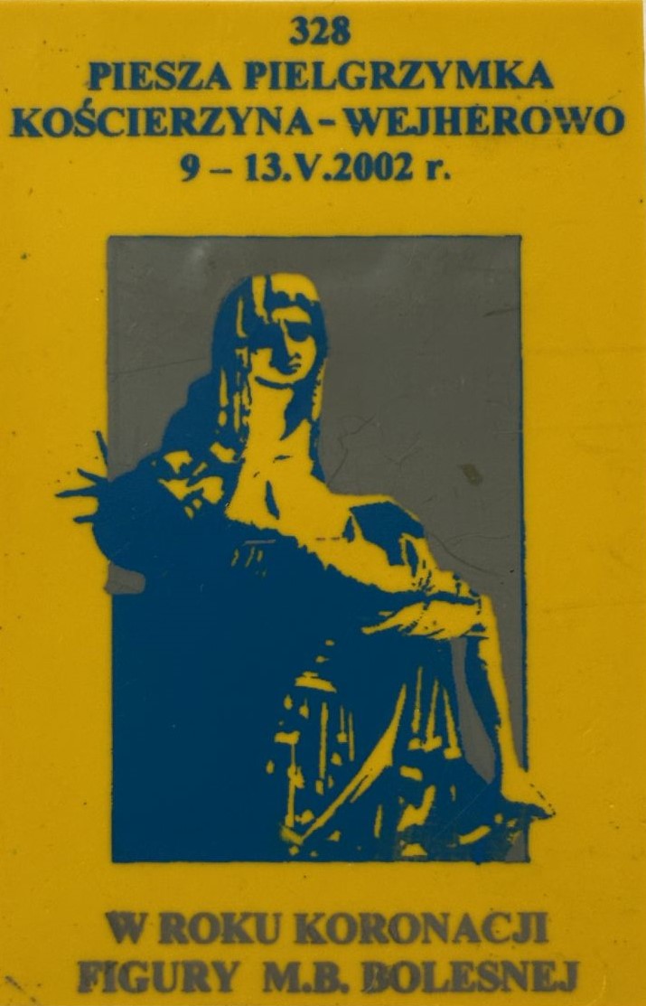 Prostokątna plakietka z pielgrzymki do Wejherowa. Na plakietce na żółtym tle wizerunek Matki Boskiej z ukrzyżowanym Jezusem.