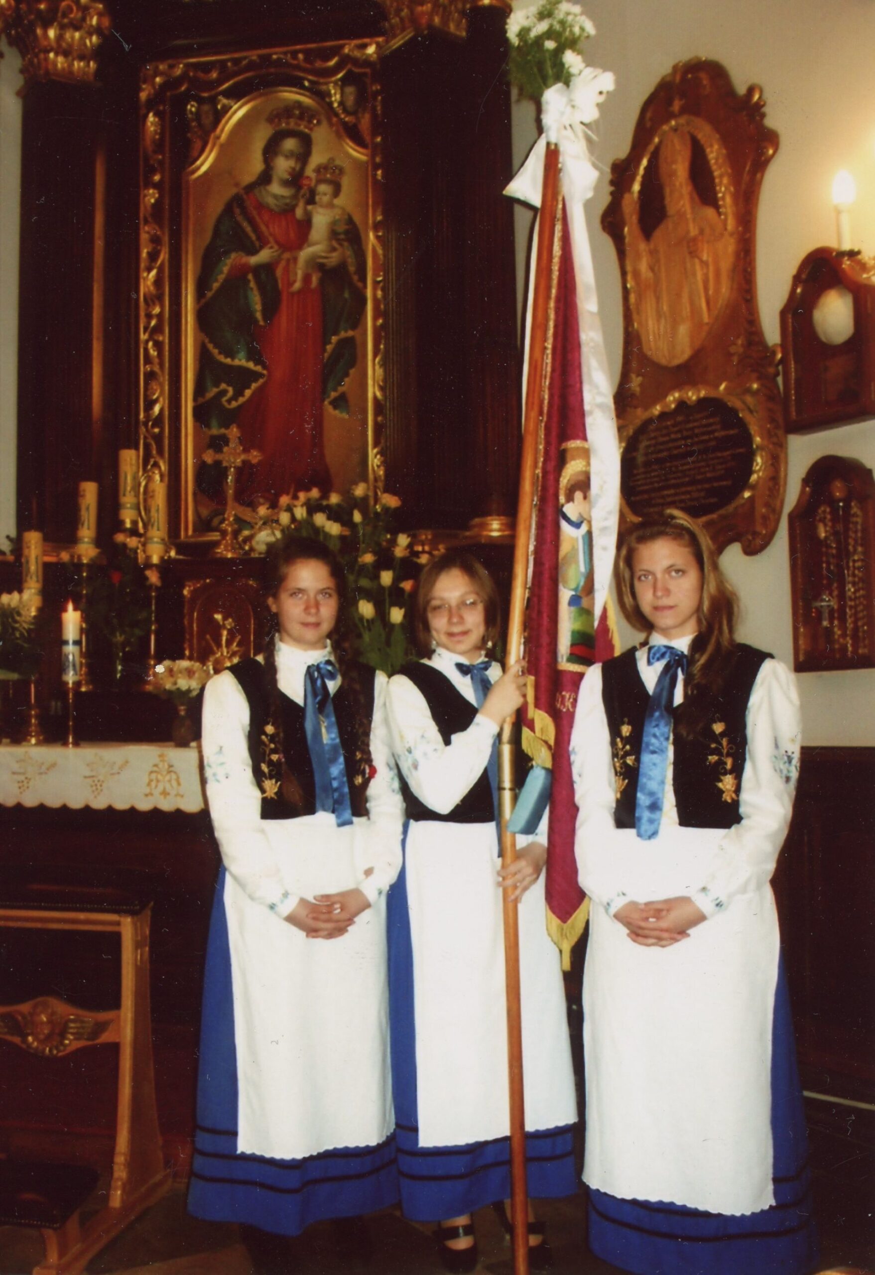 Pielgrzymka z Gdańska-Oliwy na Kalwarię Wejherowską. Trzy dziewczyny ubrane w stroje ludowe stoją w kościele z proporcem.